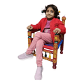 A young girl with dark hair in pigtails sits on a small, ornate, hand-painted wooden chair. She is wearing a red jacket, a pink t-shirt, pink pants, and light purple slip-on shoes.